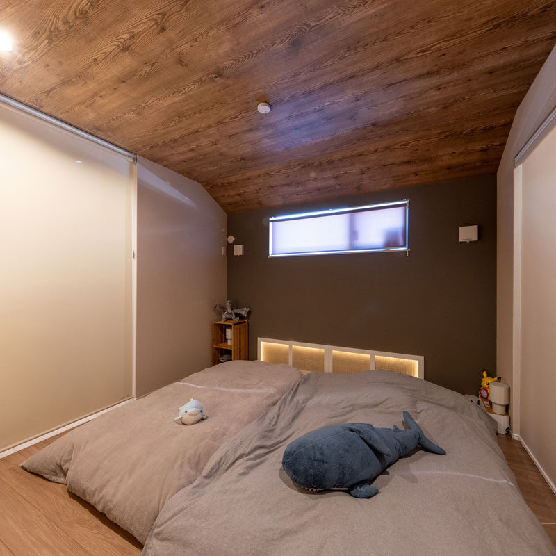 落ち着きのある寝室。クロスの色を他の居室よりワントーン落としている。照明は足元側の天井にダウンライトを、枕元にはヘッドボードの照明のような柔らかな間接照明を設置。枕元の照明はご主人のお手製で、四角に貼ったテープライトにメッシュの蓋をかぶせている。天井は、屋根の形状に合わせている。H様邸は北側斜線制限※に適合させ、北側屋根を下げた。「ロッジや別荘みたいで気に入っています」（奥様）
※北側斜線制限：北側隣地の日照を守るため、建物の高さを制限する規制